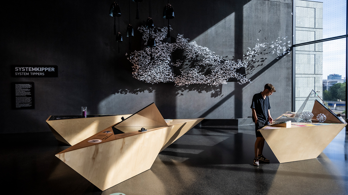 Blick in die Ausstellung mit Tischen und einer Wandinstallation aus hunderten Papiervögeln