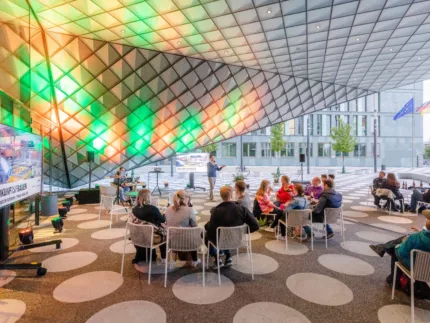 Futurium Forecourt. People are sitting around several small tables. The ceiling is illuminated by colourful lights and on the left hand side there is a tv standing in front of the wall.