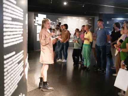 Eine Frau steht vor einer Menschengruppe. In der Hand befindet sich ein Mikrofon. Das Bild ist in der Ausstellung des Futuriums aufgenommen.