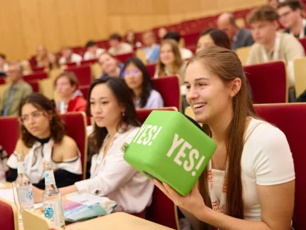 Auf dem Bild ist ein Hörsaal voller Studierender. Im Vordergrund hält eine Person einen grünen YES! Würfel.