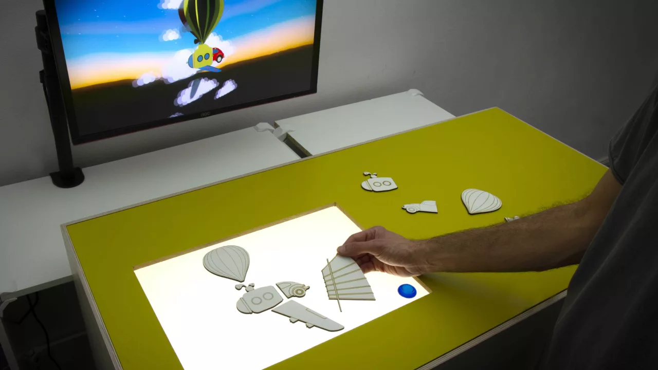 A person places punched-out parts together on a luminous surface. A screen can be seen in the background