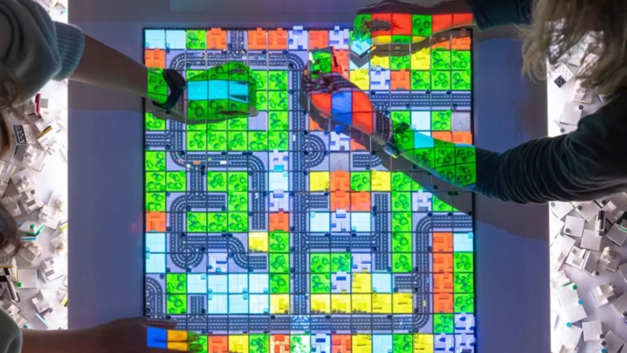 Zwei Menschen setzen bunte Klötze auf einen bunten Bildschirm. Sie bauen einen Stadtplan