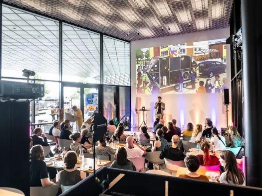 Ein Saal mit sitzendem Publikum vor einer Bühne mit Moderator, dahinter eine Projektion des Mobilen Futuriums