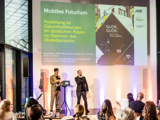 Publikum vor einer Bühne mit einem Moderator und einem Sprecher, im Hintergrund eine Projektion zur Beforschung des Mobilen Futuriums