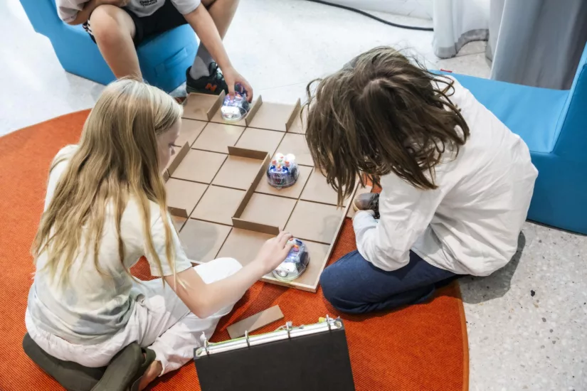 Drei Kinder sitzen vor einem Spielbrett mit Minirobotern