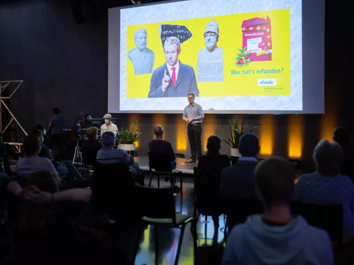 Eine Person steht auf der Bühne vor einer großen Leinwand, auf der eine Collage u. A. aus Statuen zu sehen ist. Vor der Bühne sitzt ein Publikum.