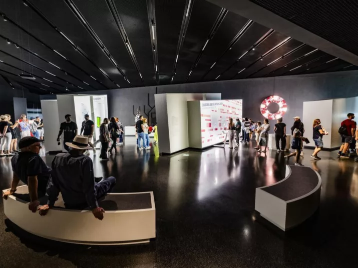 Ein Blick in den Denkraum Technik. Im Hintergrund sind Wände mit Ausstellungstehemen zu sehen. Im Vordergrund sitzen Menschen auf Bänken.