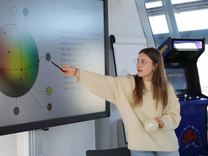 Ein junge Frau trägt einen gelben Pullover. Mit einem Stift zeigt sie auf eine Grafik. Die Grafik befindet sich auf einer digitalen Tafel.