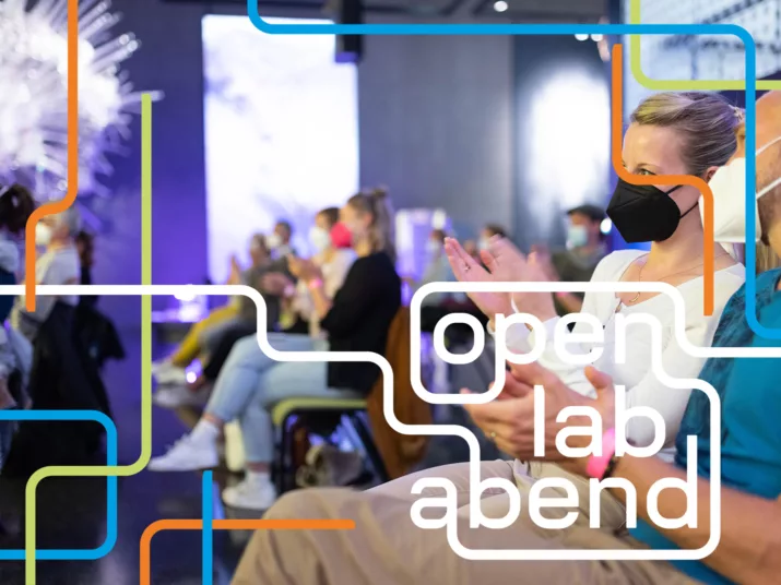 Personen sitzen im Publikum und applaudieren. Das Bild hat das Open Lab Abend-Branding.
