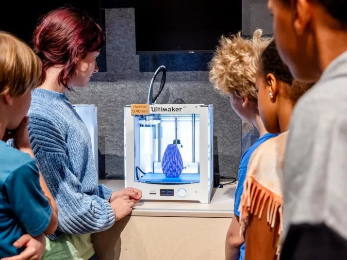 Jugendliche und Erwachsene blicken auf einen 3D-Drucker