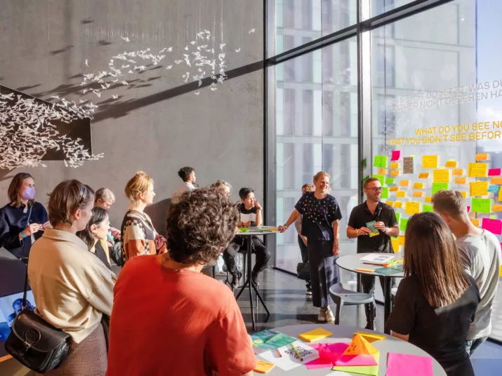Futurium Ausstellung an der großen Glaswand. Eine Gruppe steht im Kreis und diskutiert. An der Glaswand hängen viele bunte beschriftete Zettel.